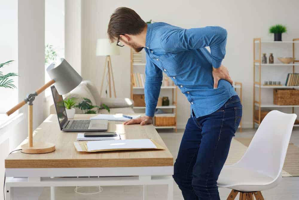 A man in a blue shirt and jeans stands up from a desk, holding his lower back with a pained expression. The workspace, featuring a laptop, notepad, and lamp, is brightened by natural light. Perhaps it's time to consider pain management NYC for relief and improved well-being.