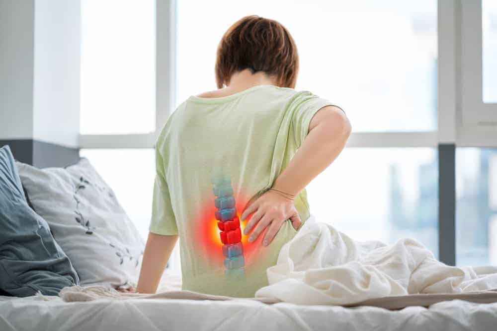 A person sitting on a bed holds their lower back, experiencing pain. The back area is highlighted with a red glow, and an illustration of the spine suggests discomfort. In this bright, softly lit NYC bedroom, they consider physical therapy for effective pain management.