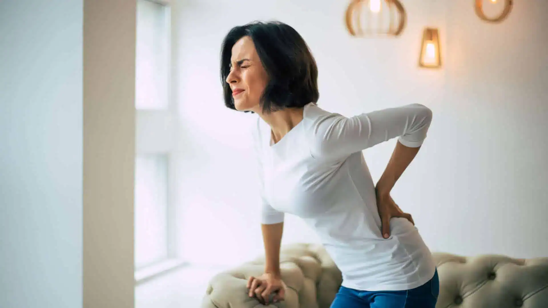 A woman with short dark hair leans on a couch by a window, wearing a white long-sleeve shirt and blue jeans. She holds her lower back with one hand, her pained expression hinting at discomfort. Considering physical therapy NYC could be beneficial for effective pain management NYC offers.