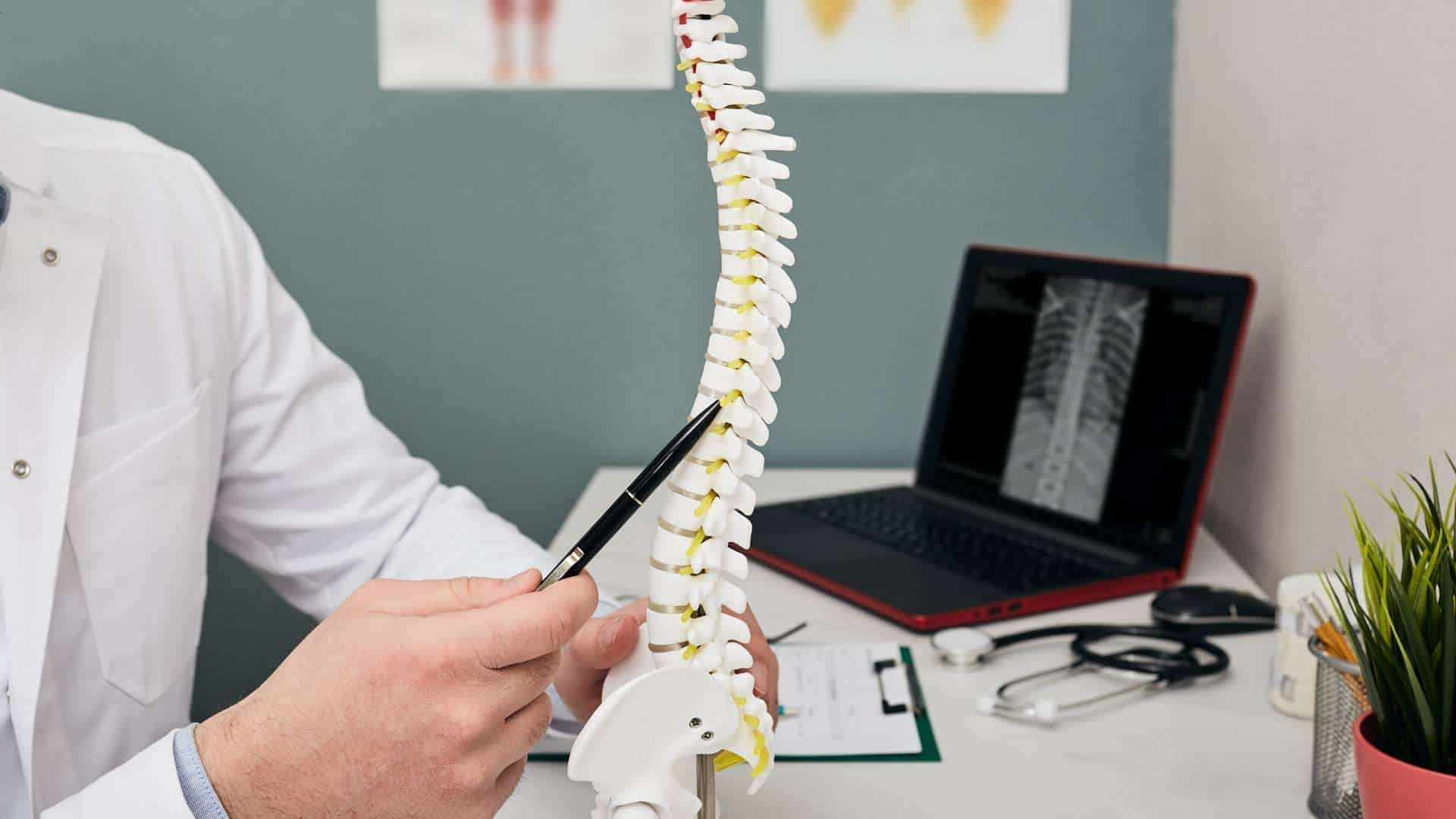 A doctor in a white coat points at a spine model with a pen while discussing pain management NYC. A laptop displays an X-ray image, surrounded by medical tools and documents on the desk.