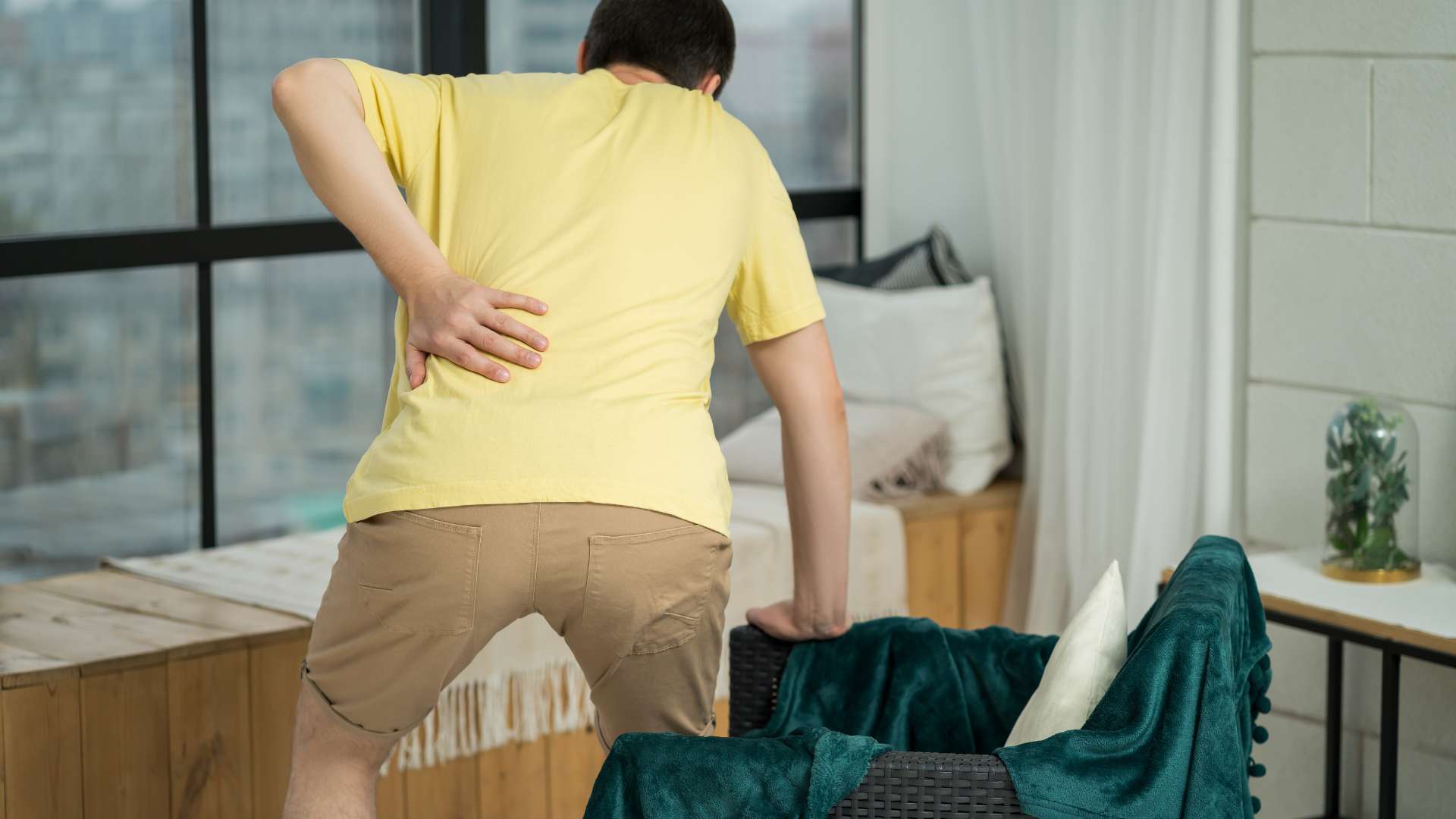 A person in a yellow shirt and brown shorts is indoors, holding their lower back with one hand while leaning on a chair, perhaps considering physical therapy NYC for relief. They appear to be experiencing back pain in a room with a window and a seating area with pillows.