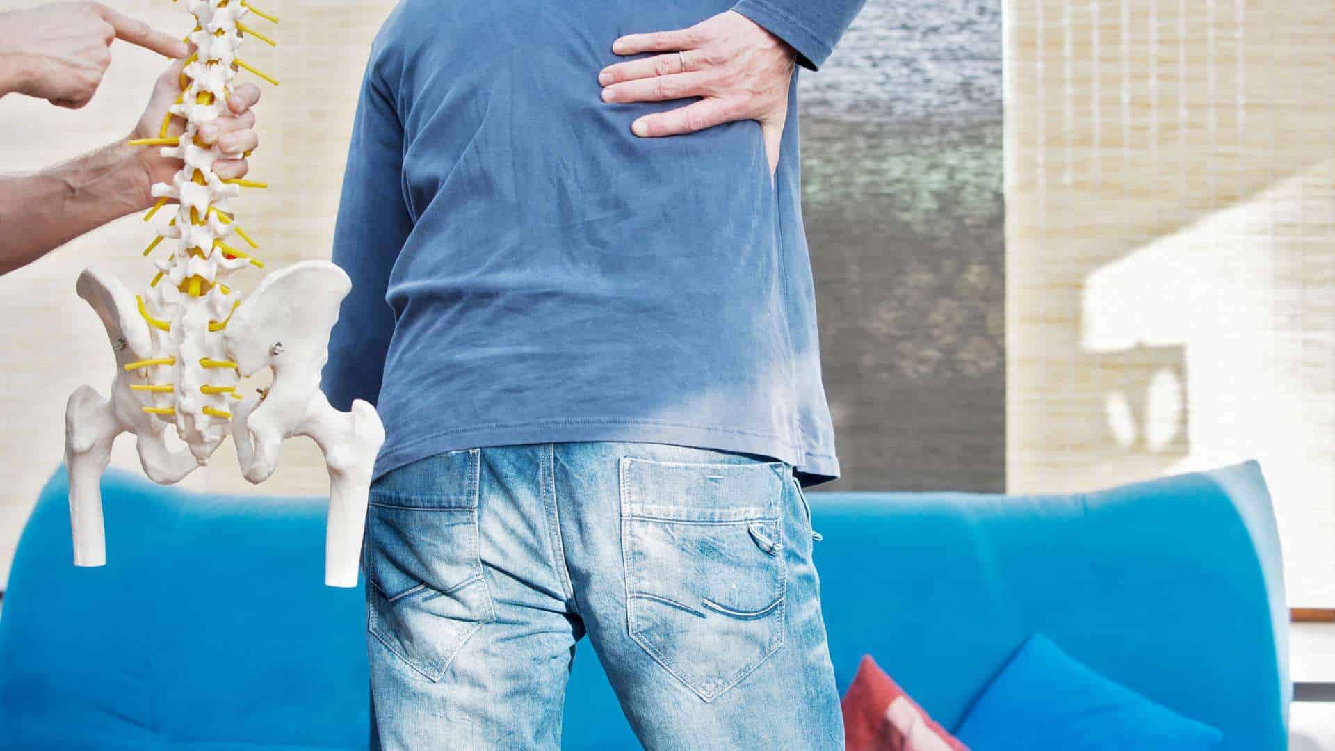 A person in a blue shirt and jeans clutches their lower back in pain as another points at a spine model, illustrating physical therapy NYC. A blue couch adorned with colorful pillows sets the backdrop.
