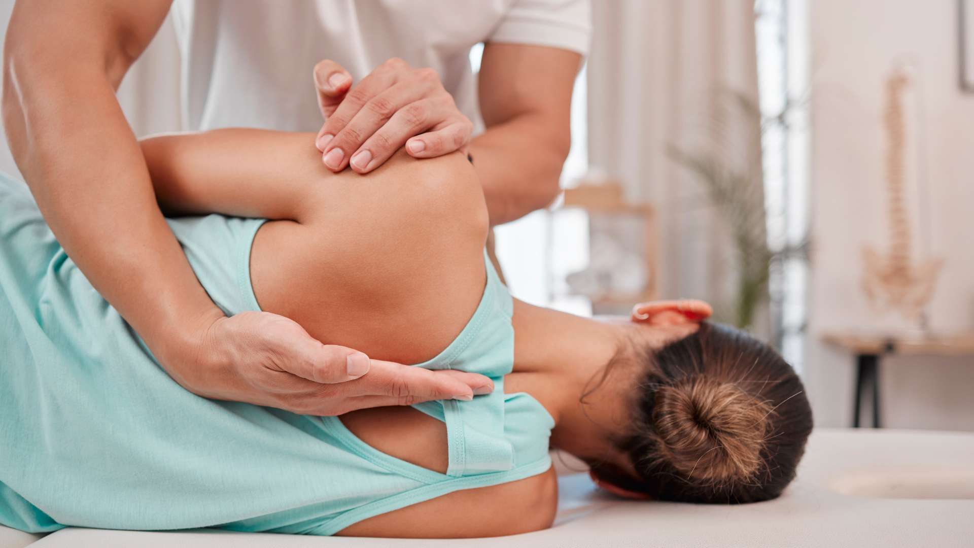 A person receiving a chiropractic adjustment while lying on their side, incorporating pain management NYC. Their arms are crossed over their chest as the chiropractor presses on their shoulder. The person, in a turquoise tank top, enjoys the bright and airy room ambiance.