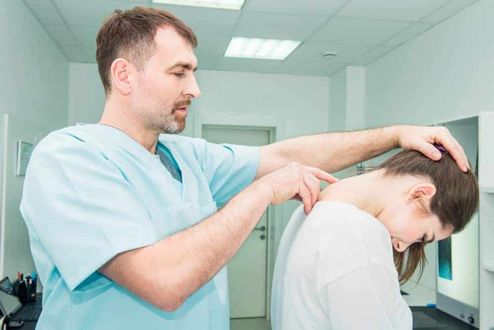 A healthcare professional in light blue scrubs examines a patient's neck, focusing on pain management NYC. The patient, with long brown hair, sits looking down as the professional gently supports their head and neck. The bright, sterile room underscores the clinical setting focused on healing.