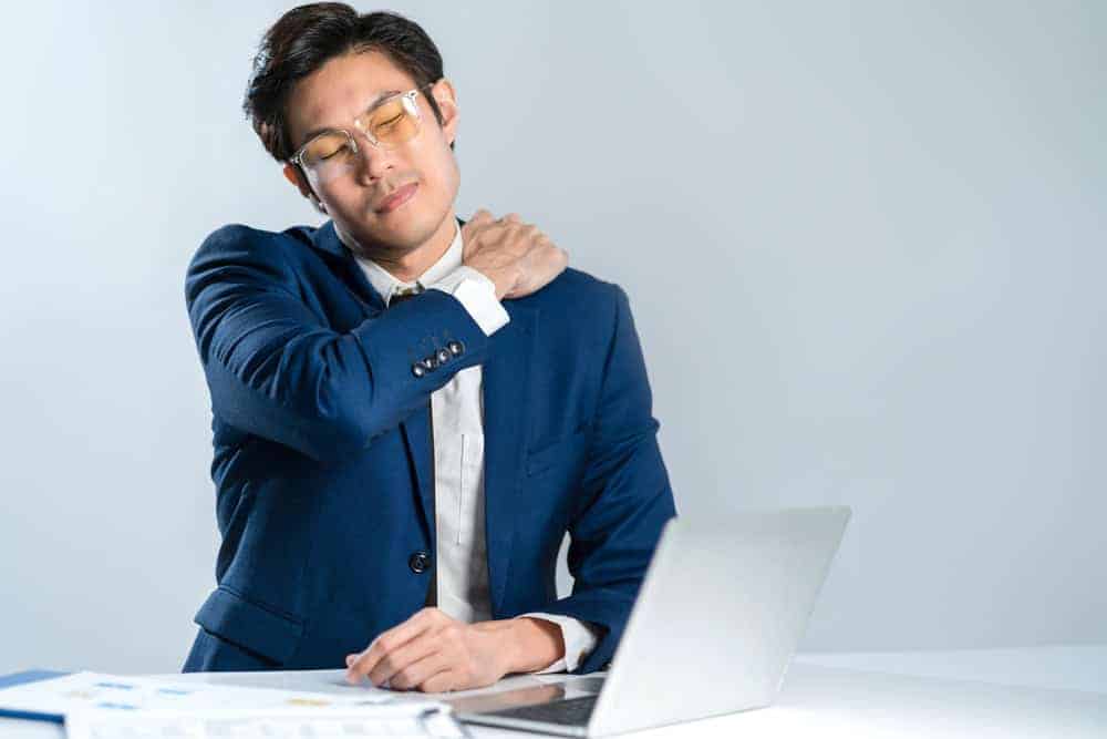 A person in a blue suit sits at a desk with a laptop, massaging their neck with a pained expression. Papers are spread out on the desk, hinting at their busy schedule. In need of some pain management or physical therapy solutions in NYC to relieve the tension.