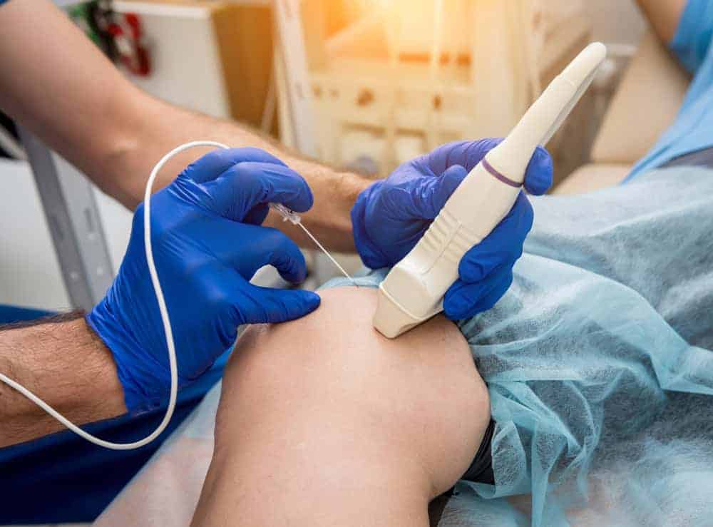 A medical professional wearing blue gloves uses an ultrasound device on a patient's knee, covered with a blue drape. The setup suggests pain management NYC procedures in progress, possibly in a clinical setting.