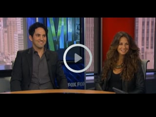 A man and a woman are sitting at a news studio desk, smiling. The backdrop features a cityscape with the words "FOX FOX," while a play button symbol floats in the center, inviting viewers to explore topics like physical therapy NYC and pain management NYC.