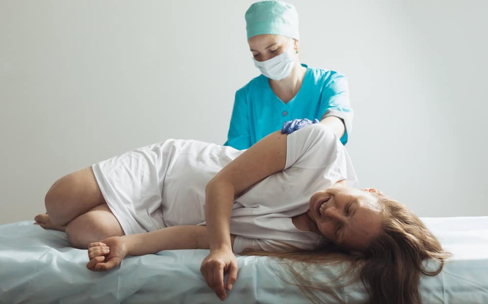 A woman in visible discomfort lies on her side during a pain management session in NYC, while a masked healthcare professional gently supports her, reflecting compassionate and expert care.
