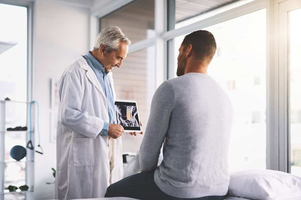 A doctor in a white coat shows an X-ray to a patient sitting on a medical examination bed, discussing pain management options in NYC. The bright room with large windows enhances their engaged conversation, reflecting the dedication found in top physical therapy practices across the city.