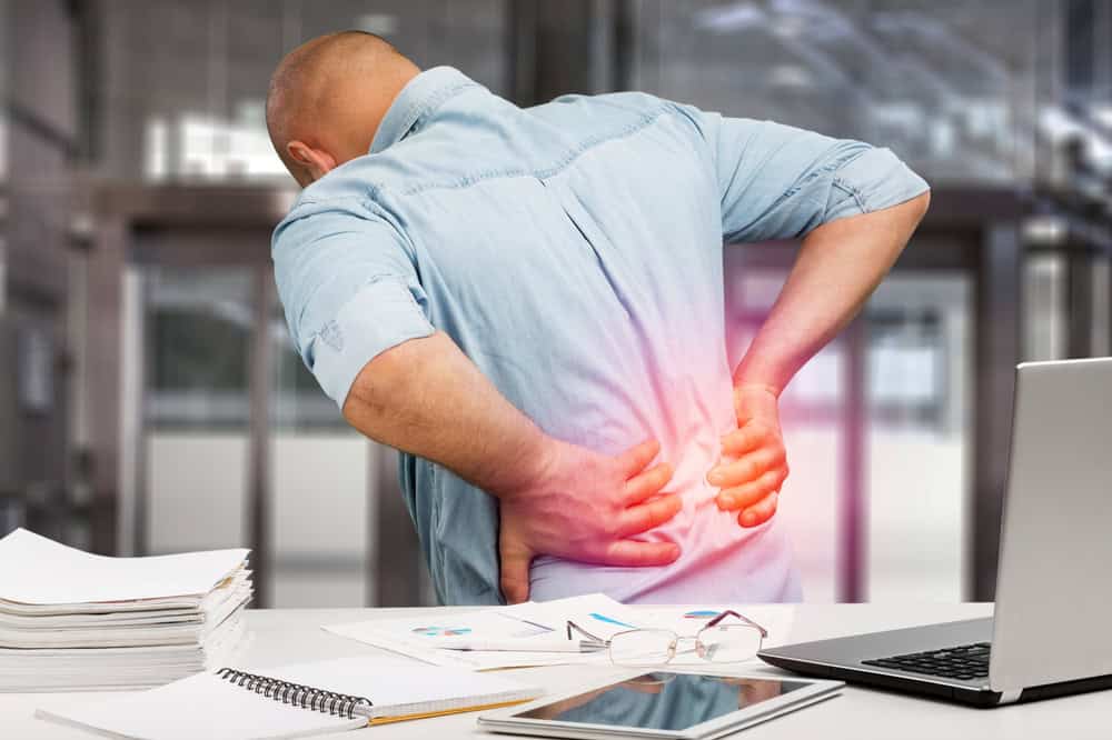 A man in a light blue shirt clutches his lower back in pain, standing beside a desk with office supplies, including a laptop, papers, and glasses. Considering physical therapy NYC might be his next step against the blurred office background.