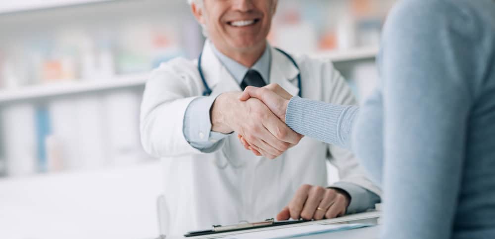 A doctor in a white coat with a stethoscope shakes hands with a patient, underscoring the importance of pain management NYC. The setting appears to be a medical office with shelves and books in the background, suggesting a friendly and professional interaction.