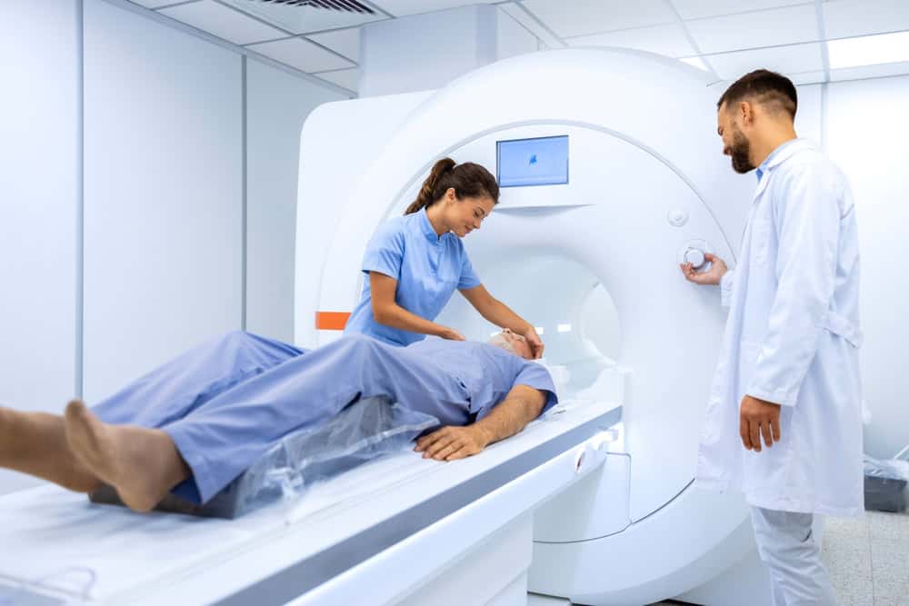 A patient lies on an MRI scanner bed, while a healthcare worker in blue assists, adjusting equipment. Another medical professional in a white coat stands beside the machine, observing. This bright, modern facility also specializes in pain management NYC services.