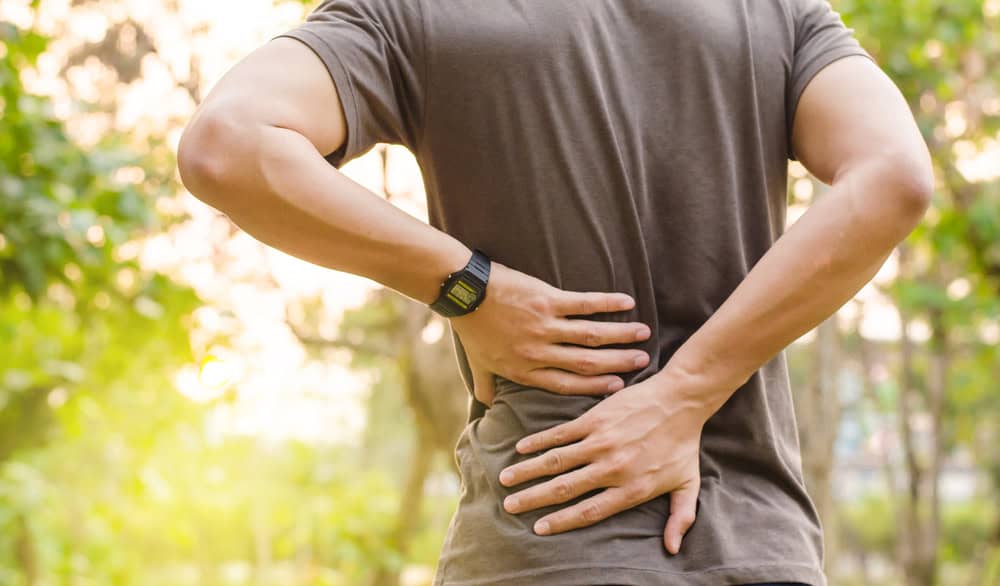 A person stands outdoors, wearing a brown shirt and black watch, holding their lower back and side with both hands, indicating discomfort. Perhaps they could benefit from pain management NYC services or physical therapy NYC amidst the blurred greenery backdrop.