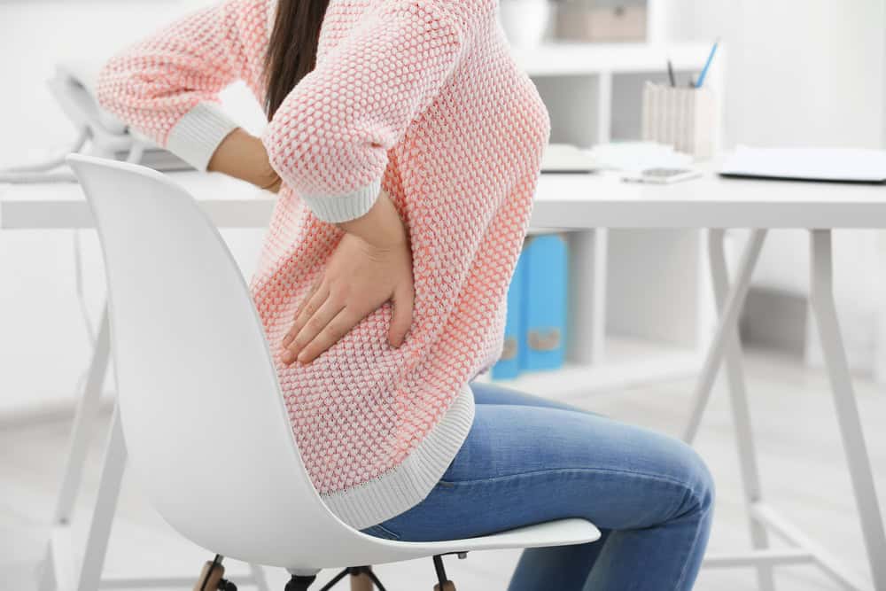 A person sits on a white chair, wearing a pink sweater and blue jeans, holding their lower back in apparent discomfort. With office supplies on the desk nearby, they seem to consider physical therapy in NYC for effective pain management.