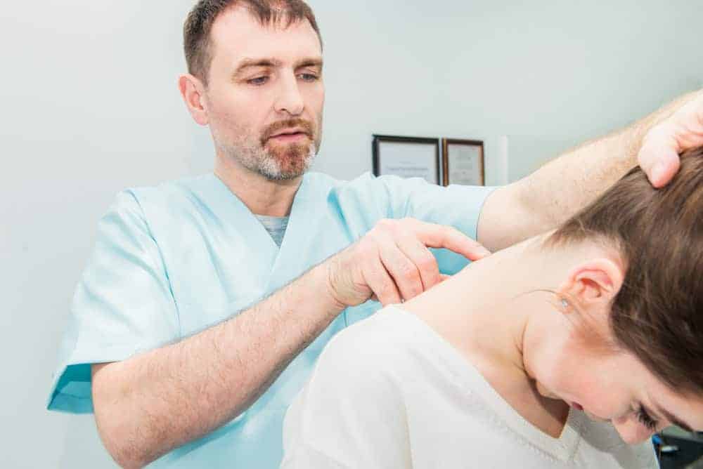 A healthcare professional in a light blue uniform examines a woman's neck and upper back, gently adjusting her posture for better pain management NYC. The woman leans forward in this clinical setting, adorned with framed certificates on the wall.