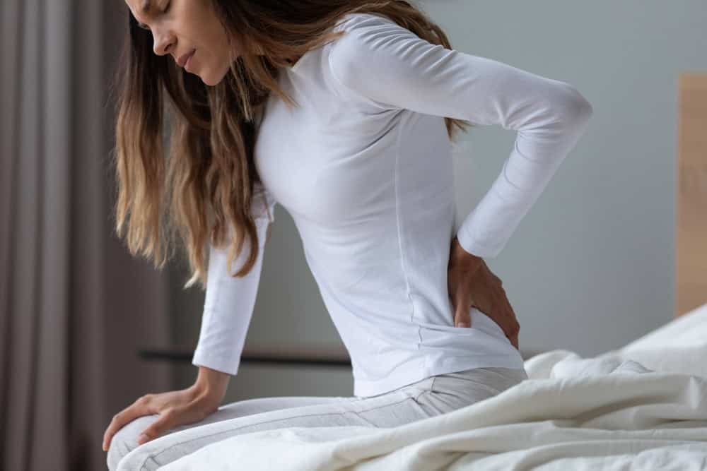 A woman sits on a bed in a brightly lit, minimally decorated bedroom, wearing a white long-sleeve top and light pants. With a pained expression, she holds her lower back, perhaps contemplating the benefits of pain management NYC to ease her discomfort.