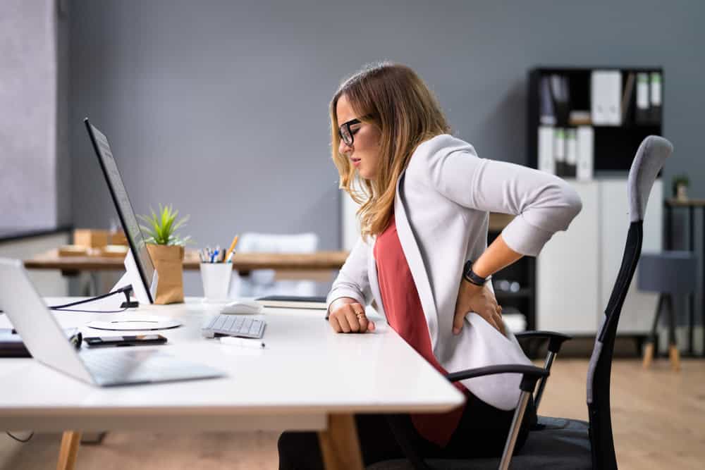 A woman sits at a desk in an office, holding her lower back with a pained expression. In her glasses, white blazer, and red shirt, she looks like she could benefit from pain management NYC services. The desk features a computer, a potted plant, and various office supplies.