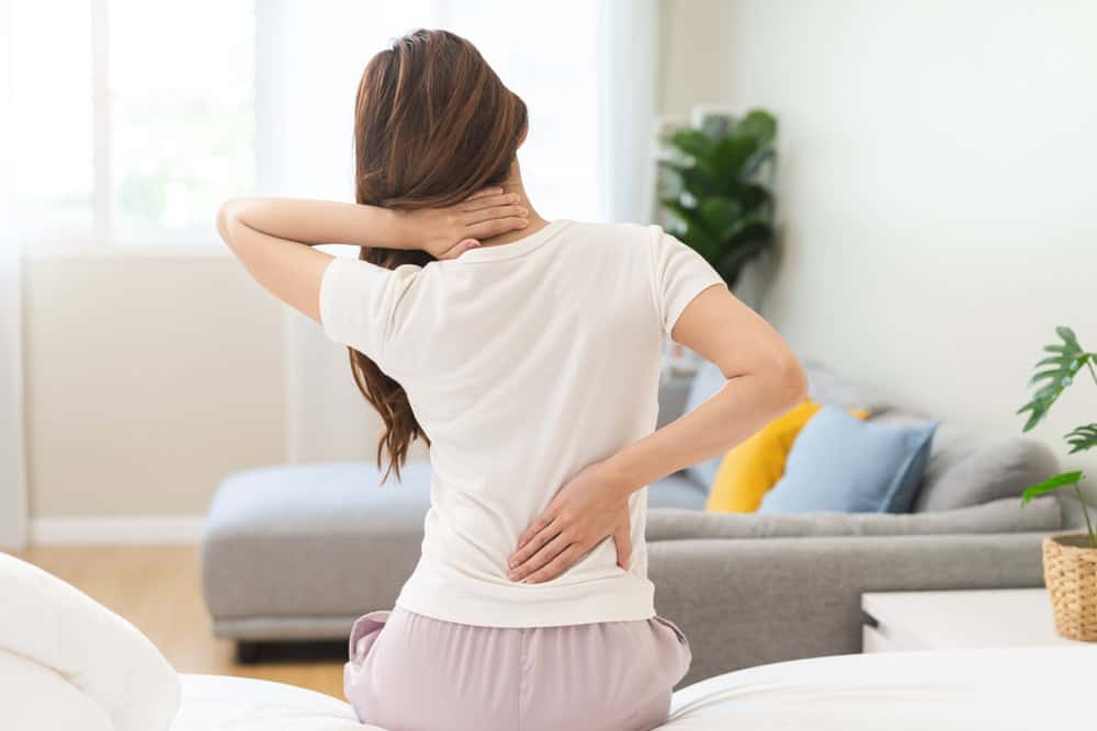A person sitting on a bed holds their neck with one hand and their lower back with the other, suggesting discomfort. They wear a white t-shirt and light pink pants. A sofa and plant are visible in the softly lit room, hinting at the need for pain management NYC solutions.