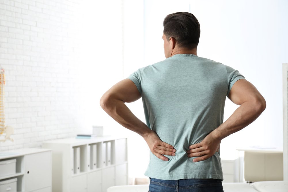 A man in a light gray t-shirt and jeans stands with his back to the camera, holding his lower back, perhaps contemplating physical therapy NYC as he seeks relief in a bright room with white walls and shelves.