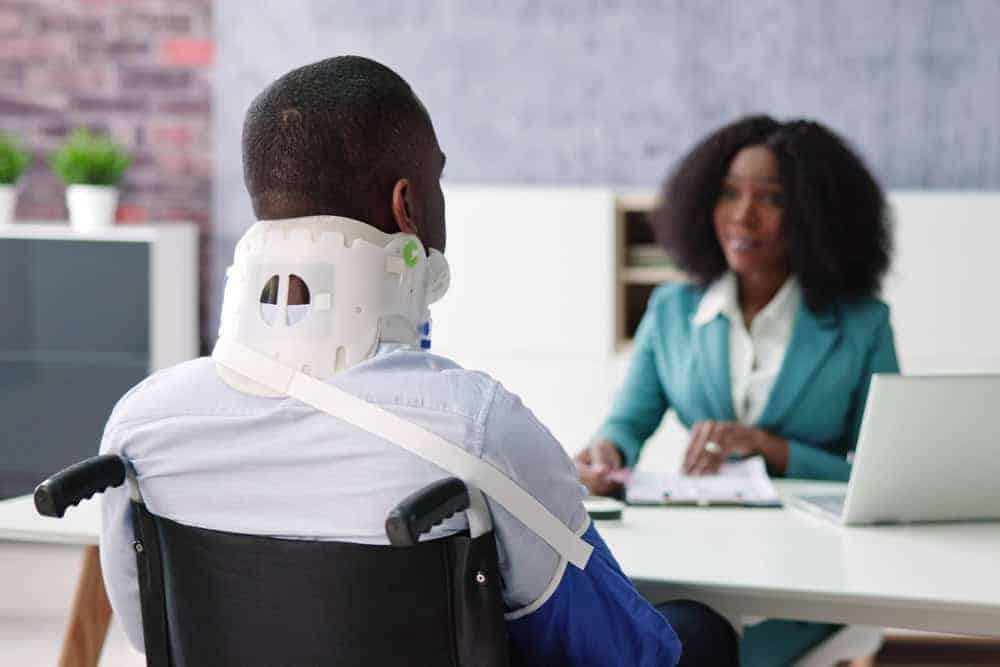 A man in a wheelchair with a neck brace sits across from a woman in a teal blazer. She is taking notes at a desk with a laptop, discussing pain management NYC solutions. They appear to be having an insightful conversation in an office setting focused on physical therapy NYC.