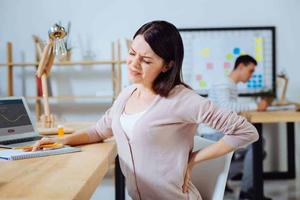 A woman sitting at a desk in an office holds her lower back with a pained expression, perhaps considering pain management NYC might offer. A laptop and a notebook are in front of her, while in the background, a person works at another desk by a colorful board.