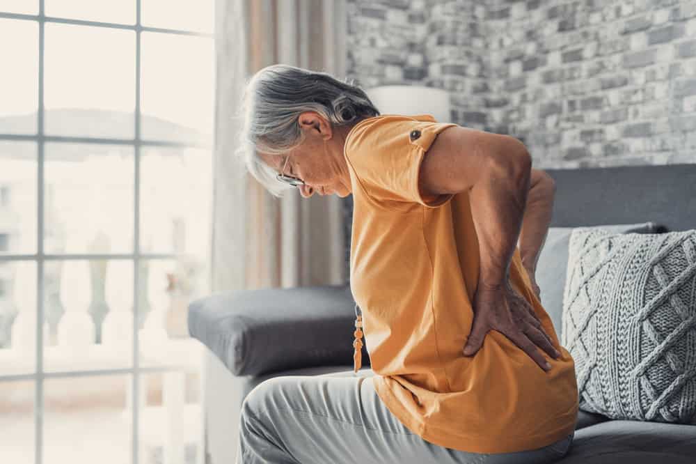 An elderly person in an orange shirt sits on a sofa, holding their lower back in discomfort. Against a backdrop of sheer-curtained windows and decorative pillows, they consider contacting top experts in pain management NYC for relief and guidance.