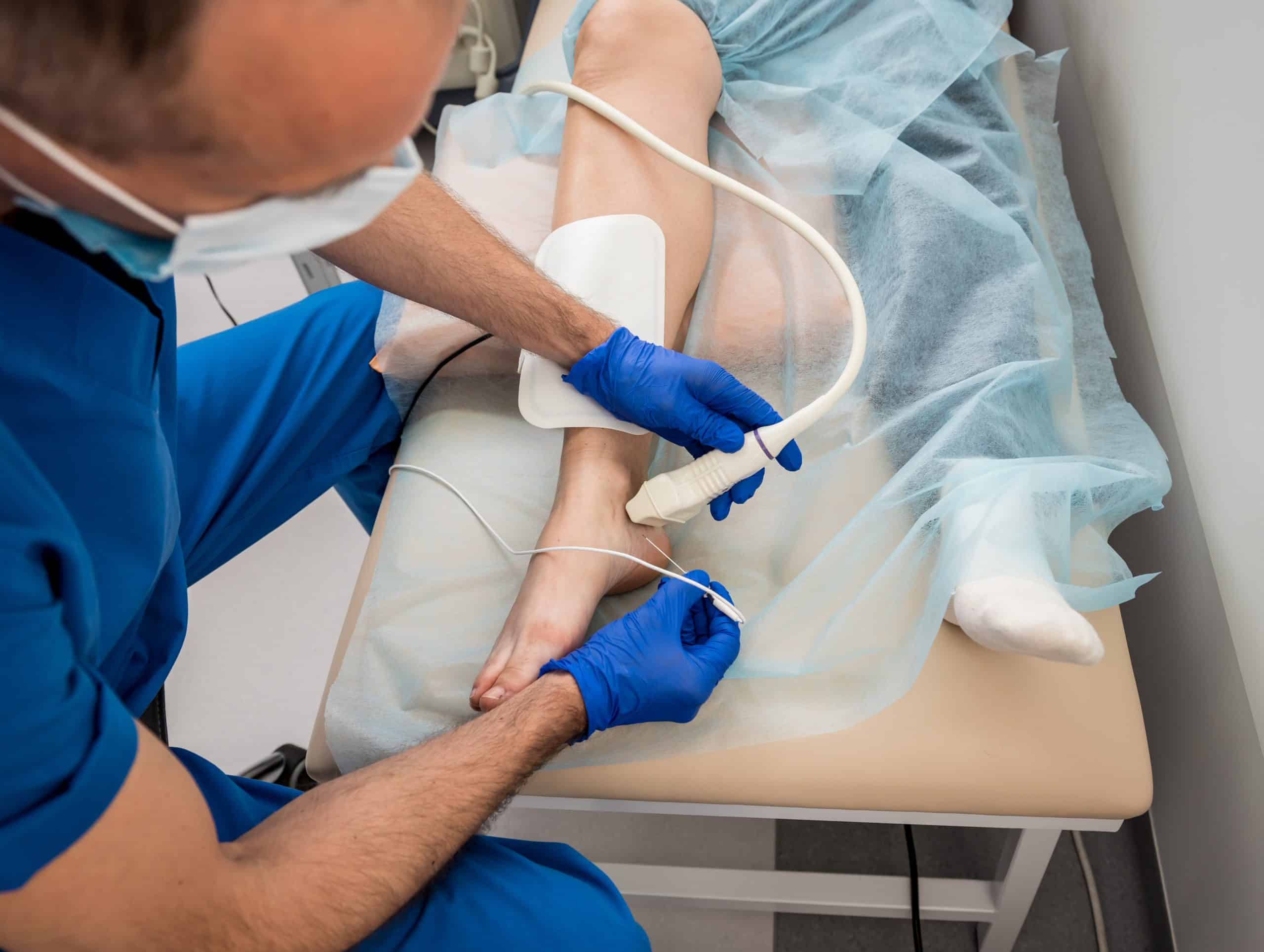 A healthcare professional in blue scrubs and gloves uses an ultrasound device on a patient's ankle, providing precise pain management in NYC. The patient lies on a bed, partially covered with a blue drape, as the focus remains on the leg and foot being examined.