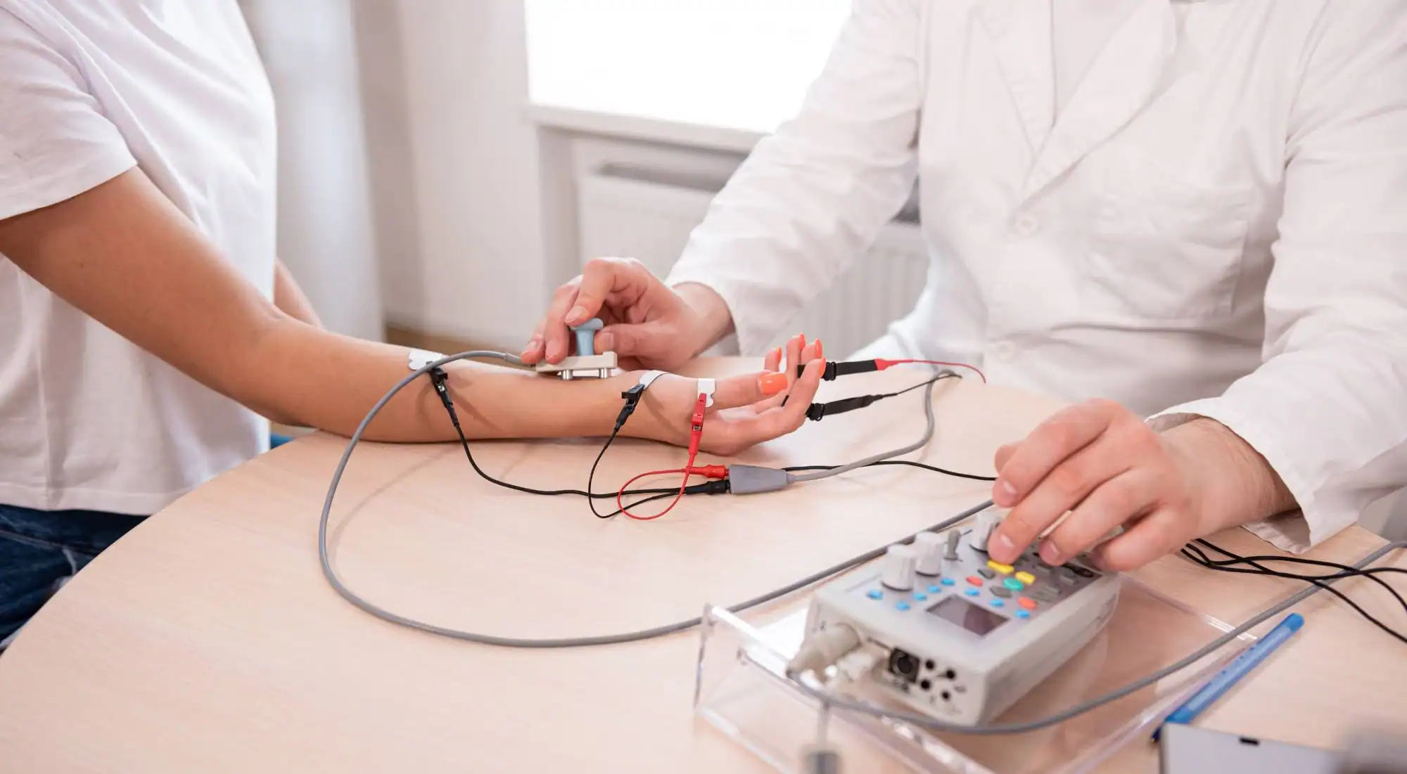 A healthcare professional attaches electrodes to a patient's fingers and wrist while operating a medical device on a table, possibly for a nerve conduction or electrophysiology test.
